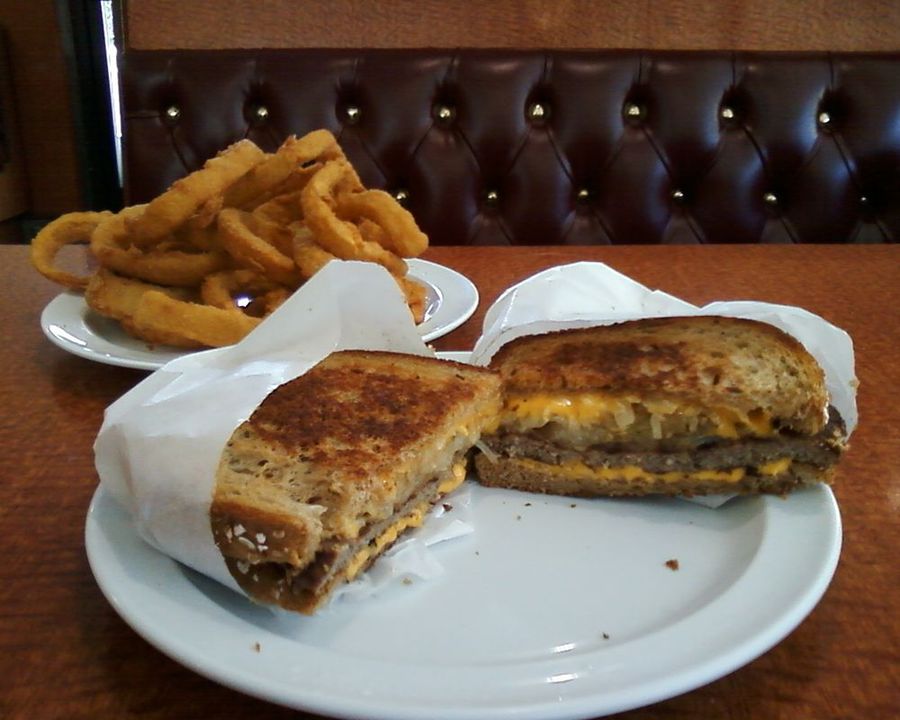 A patty melt on grilled rye with melted cheese, served with onion rings against a red tufted booth